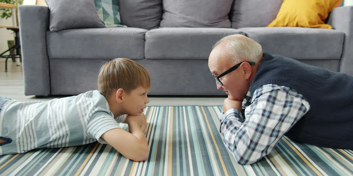 Un bambino e un uomo anziano sdraiati a pancia in giù su un tappeto a righe, uno di fronte all'altro, con il mento appoggiato sulle mani, mentre si guardano e parlano.
