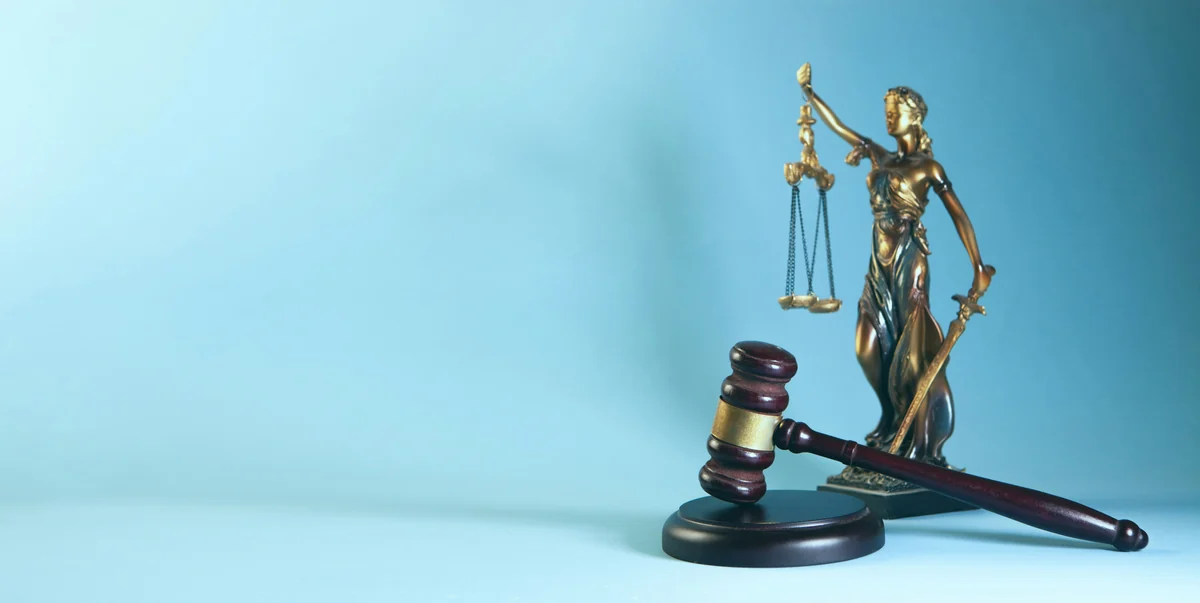 Statua della giustizia con bilancia e spada accanto a un martelletto da giudice, su fondo azzurro.